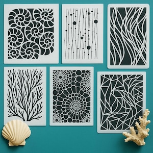 May include: Six white stencils with black designs, including swirling patterns, vertical lines with dots, and organic shapes. A seashell and coral are in the foreground. The stencils are on a teal surface.