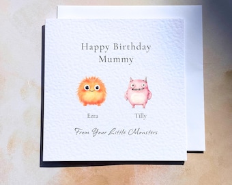 May include: A square white greeting card featuring the text Happy Birthday Mummy at the top. Below are two illustrated monsters, an orange fuzzy one labeled Ezra and a pink one labeled Tilly. The bottom text reads From Your Little Monsters.