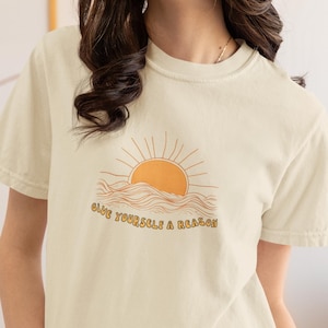 以下が含まれることがあります： クリーム色のTシャツに、海に沈む夕日のグラフィックがプリントされています。グラフィックの下には「Give yourself a reason」というテキストがプリントされています。