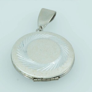 May include: A round silver-toned metal locket pendant featuring a textured, sunburst-style engraved pattern around the outer edge and a smooth, matte center. The locket includes a hinged closure at the bottom and a large, tapered bail at the top.