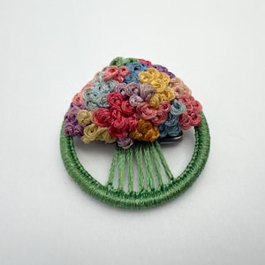 May include: A handmade brooch featuring a colorful floral design. The brooch has a green embroidered base with a circular shape and a cluster of small, colorful flowers in shades of red, pink, yellow, blue, and purple.