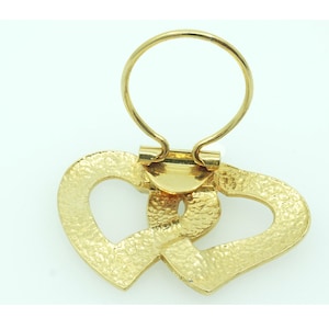 May include: A gold-toned metal clip featuring two interlocking heart-shaped charms with a textured, hammered surface finish. The accessory includes a circular ring attachment at the top, connected by a hinged mechanism to the heart-shaped base.
