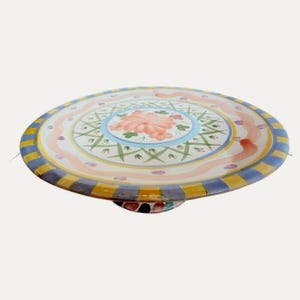 May include: A colorful, hand-painted ceramic cake stand. The round plate features a floral design with pink, blue, and green accents. The rim has yellow and blue stripes. The base is multi-colored.