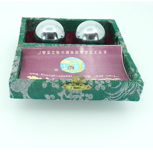 May include: Two shiny, silver metal health balls rest in a green, patterned box with a gold clasp. A card with Chinese text and a logo is visible inside the box. The box is lined with a patterned fabric.