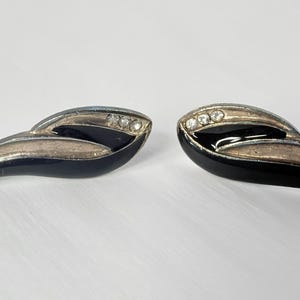 May include: A pair of vintage stud earrings. Each earring features a black enamel base with a silver-tone accent and three clear rhinestones. The earrings have a curved, leaf-like design.
