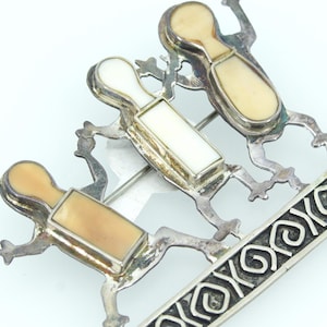 May include: A silver-toned brooch featuring three stylized figures with cream-colored and white inlays, appearing to ski on a decorative board. The brooch has a unique design with intricate details.