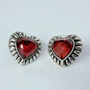 May include: A pair of heart-shaped stud earrings. Each earring features a large, faceted, red gemstone set within a silver-toned, scalloped frame. The earrings are displayed on a white surface.