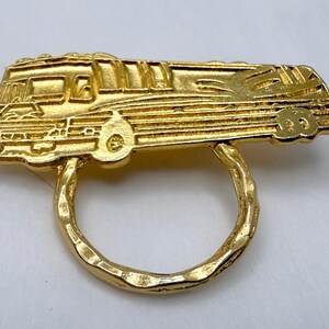 May include: A gold-coloured ring depicting a detailed recreational vehicle. The ring's design includes windows, wheels, and other features of the vehicle. The ring has a textured band.