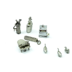 May include: A collection of small, silver-toned charms on a white background. The charms include a golf bag with clubs, a flask, a slot machine, a harmonica, a piano, and three duck figurines. The slot machine has text on it.
