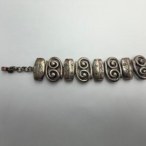 May include: Silver-toned bracelet featuring a series of decorative links. The links alternate between oval shapes with floral designs and spiral designs. The bracelet is connected by a chain and clasp.