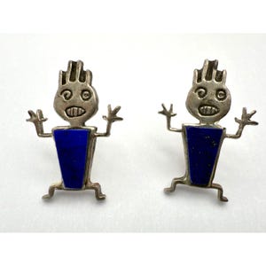 May include: A pair of silver-toned earrings featuring a stylized figure with a blue lapis lazuli body. The figures have outstretched arms and a smiling face. The earrings are designed with a Native American aesthetic.