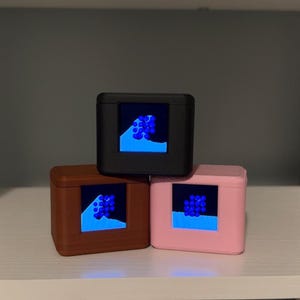 May include: Three square-shaped digital devices with matte finishes in black, brown, and light pink. Each device features a central square screen displaying a blue liquid animation with floating blue spheres against a dark background.