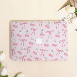 May include: A light purple laptop case featuring a repeating pattern of pink cherries and bows. The Apple logo is visible in the center. The case is designed to fit a laptop and offers a decorative and protective function.