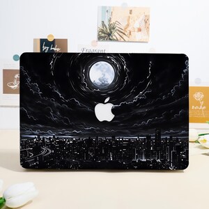 May include: A black laptop cover featuring a graphic illustration of a city skyline at night under a large, bright full moon. The design includes swirling clouds surrounding the moon and a white Apple logo cutout centered on the dark sky.