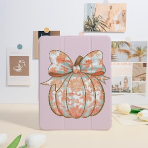 May include: A light pink tablet case featuring a pumpkin design. The pumpkin has a floral pattern in shades of orange and blue, with a gold glitter outline. The bow on top has a similar floral pattern. The case is on a white surface.