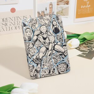May include: A black and white comic book-style case featuring Spider-Man and other characters. The case has a blue geometric design and is surrounded by other decorative items.