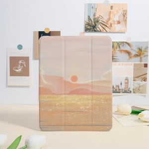 Könnte beinhalten: Eine Tablet-Hülle mit einem Sonnenuntergangs-Landschaftsdesign in Pfirsich-, Orange- und Goldtönen. Die Hülle hat ein dreifaches Design und steht aufrecht. Dekorative Postkarten befinden sich im Hintergrund.