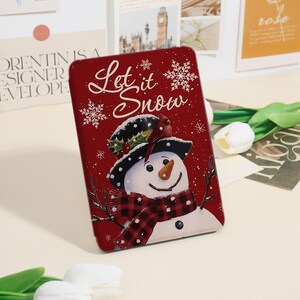 May include: A rectangular, red Christmas-themed tin featuring a snowman wearing a hat and scarf, with the words "Let it Snow" in white script. Snowflakes and a cardinal bird are also depicted.