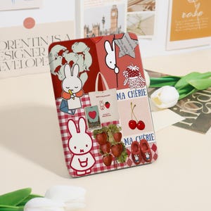 May include: A decorative case with a collage design. It features a white bunny, strawberries, cherries, and the text "MA CHERIE". The background has a red and white gingham pattern and other illustrations.