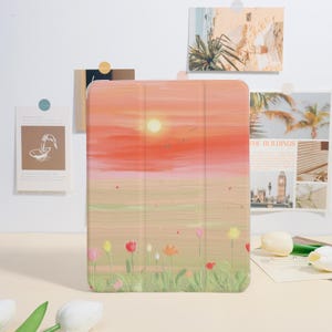 Könnte beinhalten: Eine Tablet-Hülle mit einem Sonnenuntergangs-Landschaftsdesign, das einen orange-gelben Himmel, eine helle Sonne und Vögel im Flug zeigt. Die Hülle hat auch ein Feld mit bunten Blumen am unteren Rand. Die Hülle befindet sich auf einer weißen Oberfläche.
