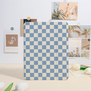 Peut inclure: Une housse de tablette à carreaux bleus et crème. L'étui rectangulaire présente un motif quadrillé de carrés bleus et crème alternés. L'étui est posé verticalement sur une surface claire.