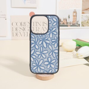 May include: A phone case with a floral design. The case has a black border and a light blue and white floral pattern. The phone case is displayed on a small wooden stand.