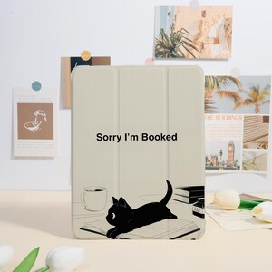 Peut inclure: Une housse de tablette beige avec le texte "Sorry I'm Booked" et une illustration de chat noir. Le chat est allongé sur un livre ouvert, avec une tasse à café et des livres en arrière-plan. La housse est sur une surface claire.