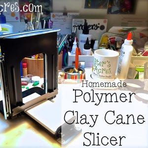 Può includere: Un taglierino per canne di argilla polimerica nero con base bianca. Il taglierino ha un telaio rettangolare e una lama per tagliare l'argilla. L'immagine include anche una tazza con la scritta "Kater's Acres" e il testo "Homemade Polymer Clay Cane Slicer."