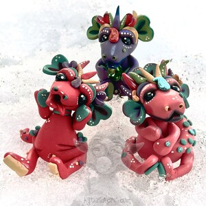 May include: Three small, hand-crafted, colorful dragons made of polymer clay. The dragons are sitting on a white surface. One dragon is purple with green and yellow accents. The other two dragons are red with green, yellow, and white accents.