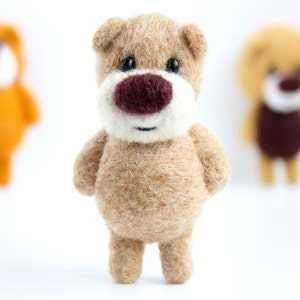 May include: A brown felt teddy bear with a white face and a brown nose. The bear is standing on a white surface.