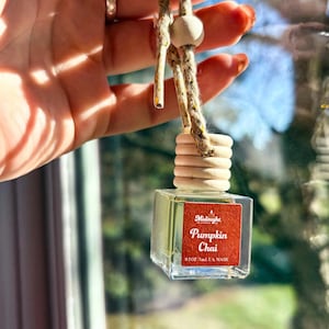 May include: A small, clear glass bottle with a wooden cap and a rope hanger. The bottle has a red label with the words "Pumpkin Chai". The bottle contains a liquid, and is suspended in front of a window.