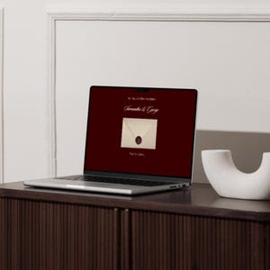 May include: A laptop screen shows a burgundy background with the text "Samantha & George" above an envelope image with a wax seal. The laptop is on a dark wood cabinet, with a white decorative object to the right.