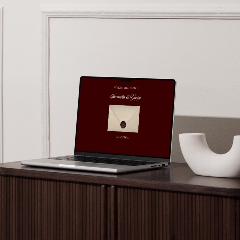May include: A laptop screen shows a burgundy background with the text "Samantha & George" above an envelope image with a wax seal. The laptop is on a dark wood cabinet, with a white decorative object to the right.