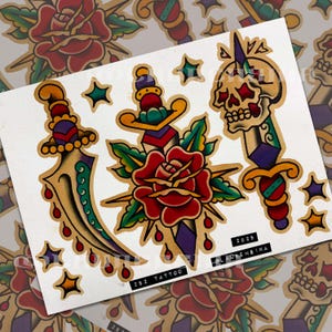 May include: A tattoo flash sheet featuring traditional style illustrations of a curved dagger, a rose pierced by a dagger, and a skull pierced by a dagger, accented with stars. The text 252 TATTOO, 2025, and LAURENSIKA is printed on the sheet.