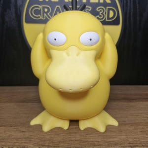 May include: A yellow duck-like Pokemon figure with its hands covering its ears. The figure is standing on a wooden surface.