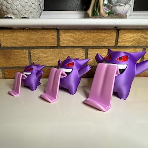 May include: Three purple Gengar-shaped phone stands with pink phone rests. The Gengar figures have red eyes and wide, toothy grins. The stands are arranged in a row, decreasing in size from right to left.