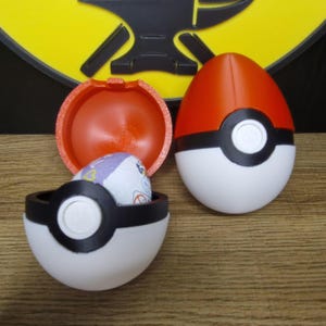 Peut inclure: Deux contenants en forme de Poké Ball rouge, blanc et noir. Un contenant est ouvert et montre un bonbon violet et blanc à l'intérieur. L'autre contenant est fermé.