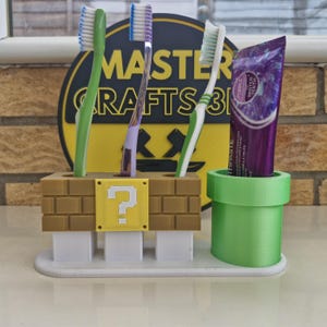 May include: A novelty toothbrush holder, 3D-printed in the style of a video game. It features a brown brick design with a yellow question mark block and a green pipe for toothpaste. Three toothbrushes and a tube of purple toothpaste are visible.