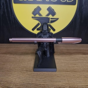 Puede incluir: Una figura negra de Darth Vader sosteniendo una pluma de color rosa dorado. La figura está arrodillada y tiene una base negra.