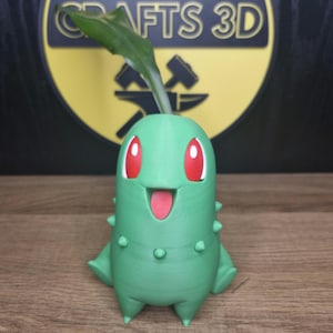 Pode incluir: Um vaso verde impresso em 3D com a forma de um personagem Pokémon, Bulbasaur, com olhos vermelhos e um sorriso largo. Uma pequena planta verde está crescendo no topo do vaso.