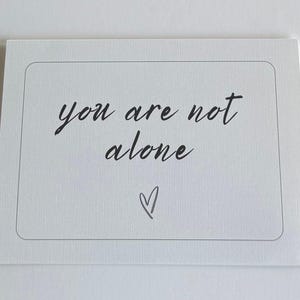 Może przedstawiać: Biała karta z zaokrąglonymi rogami i cienką szarą obwódką. Słowa "you are not alone" są napisane czarnym atramentem w stylu odręcznym. Poniżej znajduje się małe serce.