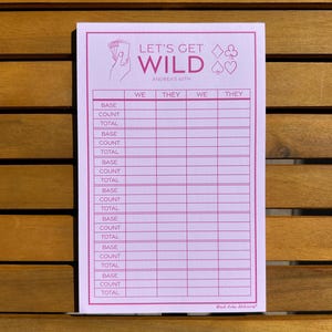 May include: A pink score sheet for a game, featuring the text "LET'S GET WILD" and "ANDREA'S 60TH". The sheet is divided into columns for "WE" and "THEY", with rows for "BASE", "COUNT", and "TOTAL". A hand holding playing cards is in the upper left corner.