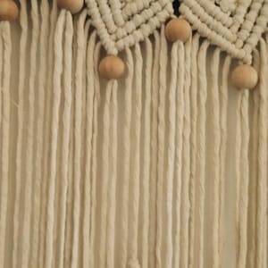 Puede incluir: Colgante de pared de macram&eacute; color crema con un dise&ntilde;o geom&eacute;trico y cuentas de madera. La pieza presenta una secci&oacute;n superior tejida con patrones de diamantes y largas franjas verticales.