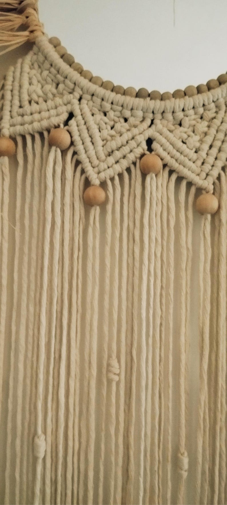 Puede incluir: Colgante de pared de macram&eacute; color crema con un dise&ntilde;o geom&eacute;trico y cuentas de madera. La pieza presenta una secci&oacute;n superior tejida con patrones de diamantes y largas franjas verticales.