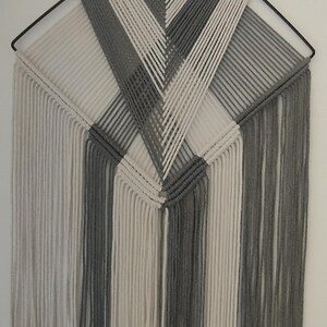 May include: A grey and white macrame wall hanging with a geometric pattern. The hanging has long fringe at the bottom.