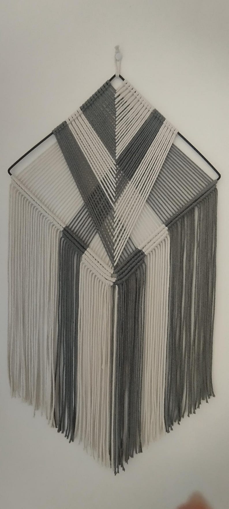 May include: A grey and white macrame wall hanging with a geometric pattern. The hanging has long fringe at the bottom.