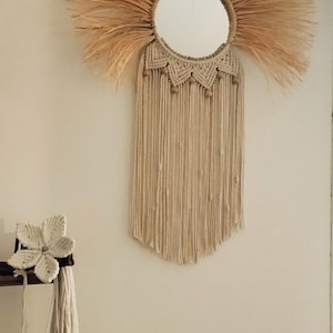 Può includere: Uno specchio rotondo con un design a raggiera in fibra naturale. Lo specchio è circondato da un materiale intrecciato simile alla rafia, con una frangia in stile macramè sottostante. Decorazione murale in stile bohémien.