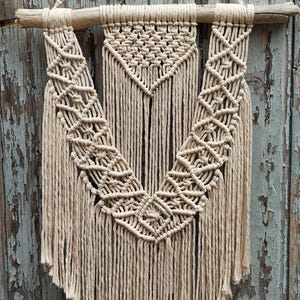 Peut inclure: Suspension murale en macramé beige avec un motif géométrique et des franges. La décoration murale est suspendue à une cheville en bois naturel et à une corde tissée. Le fond est un mur en bois bleu clair vieilli.