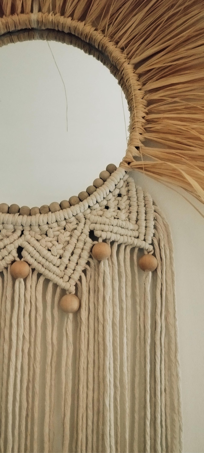 Puede incluir: Un espejo redondo con un marco de fibra natural y detalles de macram&eacute;. El marco est&aacute; hecho de un material tejido similar a la paja. El macram&eacute; presenta un patr&oacute;n geom&eacute;trico con cuentas de madera y flecos colgantes. La paleta de colores es neutra, con tonos beige y marr&oacute;n.