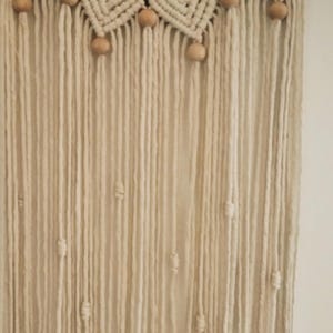 Puede incluir: Un tapiz de pared de macram&eacute; color crema con un dise&ntilde;o circular. La parte superior presenta un patr&oacute;n tejido con cuentas y detalles de madera. Largos hilos verticales de hilo cuelgan debajo, con cuentas de madera espaciadas a lo largo. Est&eacute;tica bohemia.